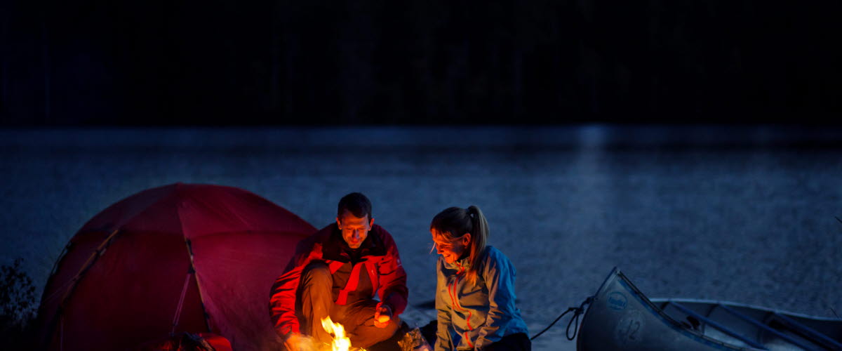 Ett par sitter vid utanför sitt tält vid en eld på kvällen. Bakom dem ser man en sjö och en skog. En kanot ligger vid sjökanten.
