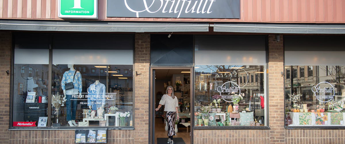 Exterior view of the Tourist Information Center. Large display windows in a brick building. In the open doorway stands Sara, who runs the tourist center.