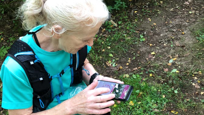 Kvinna med ljust hår i en tofs tittar ner i sin telefon och checkar in på sin kontroll i Billingen Open Trail. Bakom ser man buskar och träd.