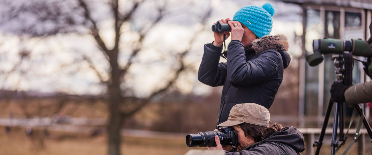 tre persomer som står vid hornborgasjön och fågelskådar med kikare och en kamera