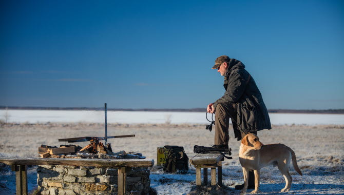 Man som står vid en grillplats med sig hund. Blå himmel i bakgrunden.