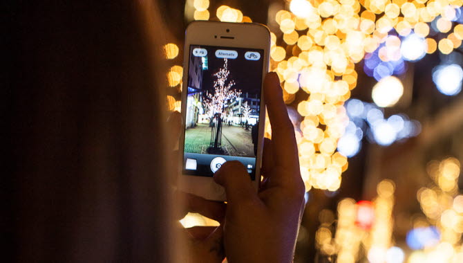 En person med långt hår håller upp en vit mobiltelefon och tar foto på ett träd med ljusslingor. Personen befinner sig utomhus och det är mörkt.