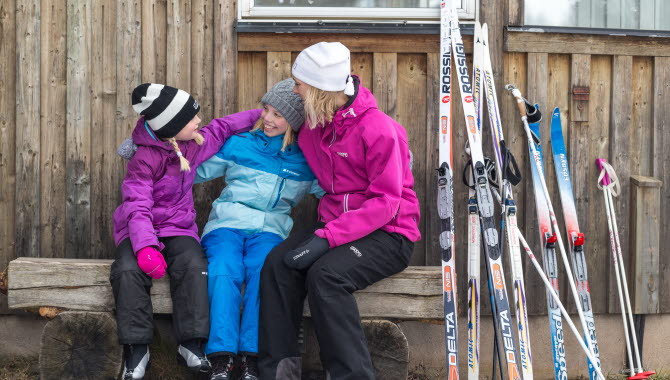 En mamma och två barn som sitter på en bänk utomhus och det står längdskidor mot väggen.