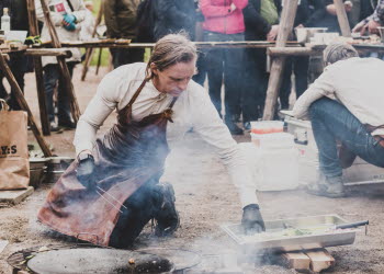 En man som lagar mat utomhus med publik bakom sig. 