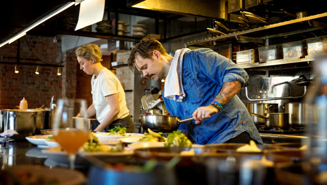 Two chef´s are cooking dinner in a restaurant. In front of them there are several plates with vegetables on it.