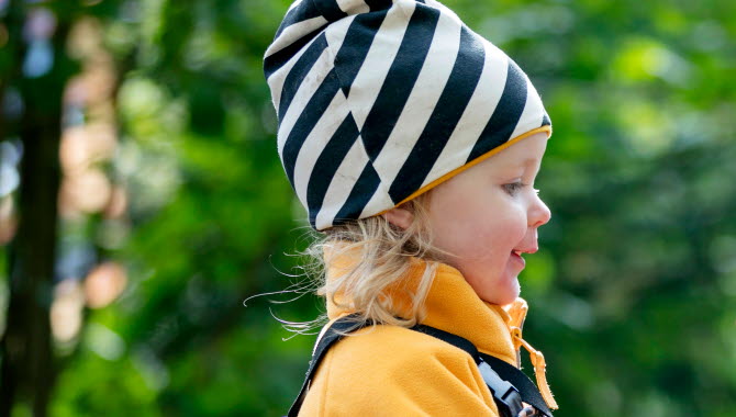 Liten blond flicka är ute i naturen. På sig har hon en randig, svart och vit mössa samt en gul fleecejacka. 