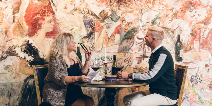 En kvinna i ljust hår och en man med kort ljust hår sitter vid ett bord med ett stort konstverk på väggen bredvid sig. 