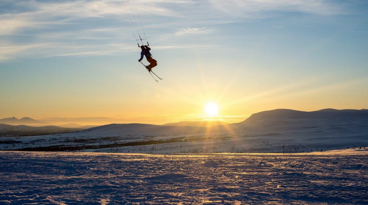 En skidåkare glider fram i luften med skidor på fötterna och fallskärm på ryggen i solnedgång.