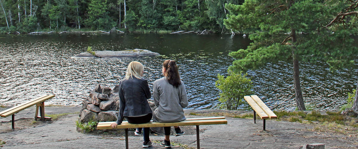 Två personer sitter på en bänk vid en grillplats på klippor vid vatten. 