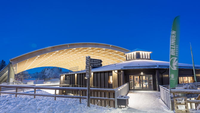 Upplyst svart byggnad(Billingecenter) i skymningen. Det liger snö på backen och i bakgrunden ser man det välvda taket över isbanan. 