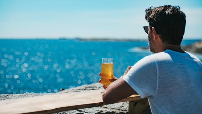 Man holding a beer, looks out over a clear blue ocean and sky.