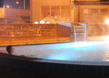 A man sits in a steaming outdoor pool. It is dark. In the background there is a wooden sauna with large windows.