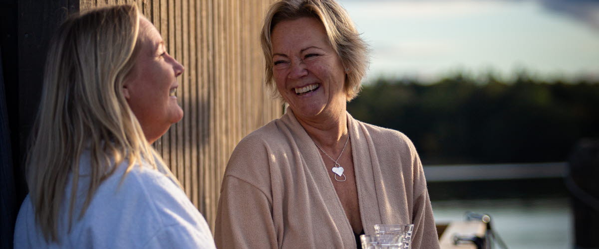 Två kvinnor pratar och skrattar med ett glas i handen på däcket av bastuflotten