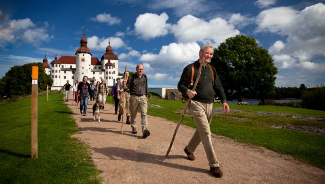 A man in the foreground and behind him, he has a line of hikers. The path goes outside the large magnificent white Läckö Castle.