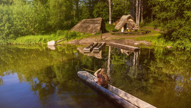 En tjej som paddlar stockbåt fram till en boplats med hyddor och eld.