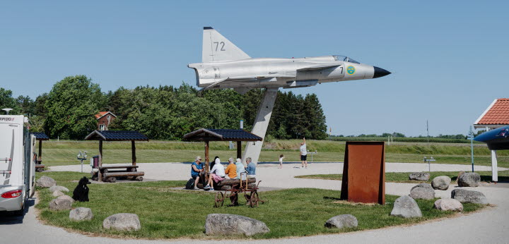 Rastplats Viggen i Grästorp med stort flygplan. Människor sitter och fikar vid ett bord. En förälder och ett barn tittar på flygplanet och pekar
