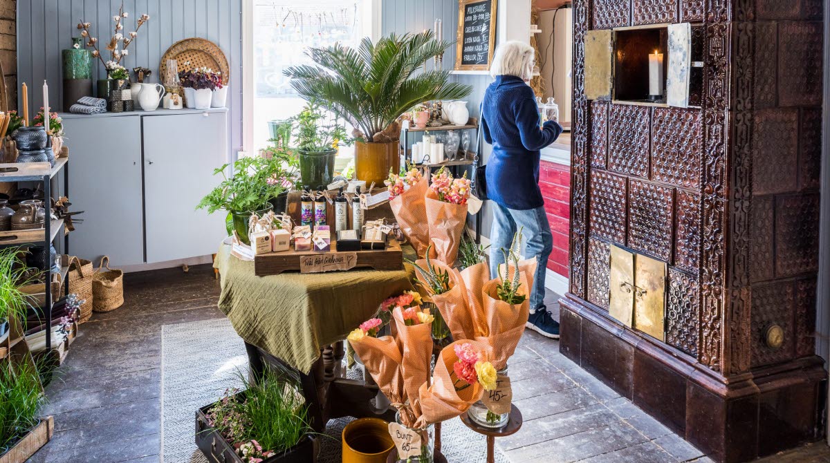 En bild inifrån en liten butik med en stor brun kakelugn. Mitt på golvet står ett bord pyntat med blombuketter och vackra tvålar.