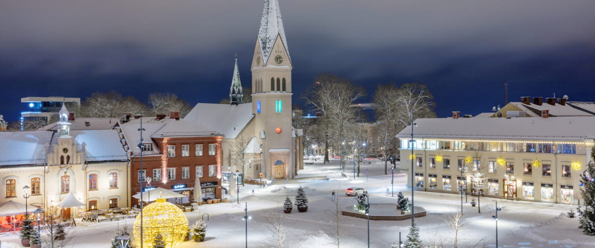 Vy över ett snötäckt Hertig Johans torg med kyrkan i bakgrunden och stämningsfull julbelysning. 
