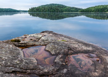 En bild på en stenklippa som ligger omgivet av vatten från en stor sjö. I bakgrunden ser man en stor grön skog.