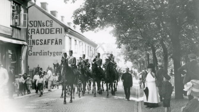 Fem husarer till häst rider fram utmed en kullerstensgata. På sidorna av gatan står människor och tittar på.
