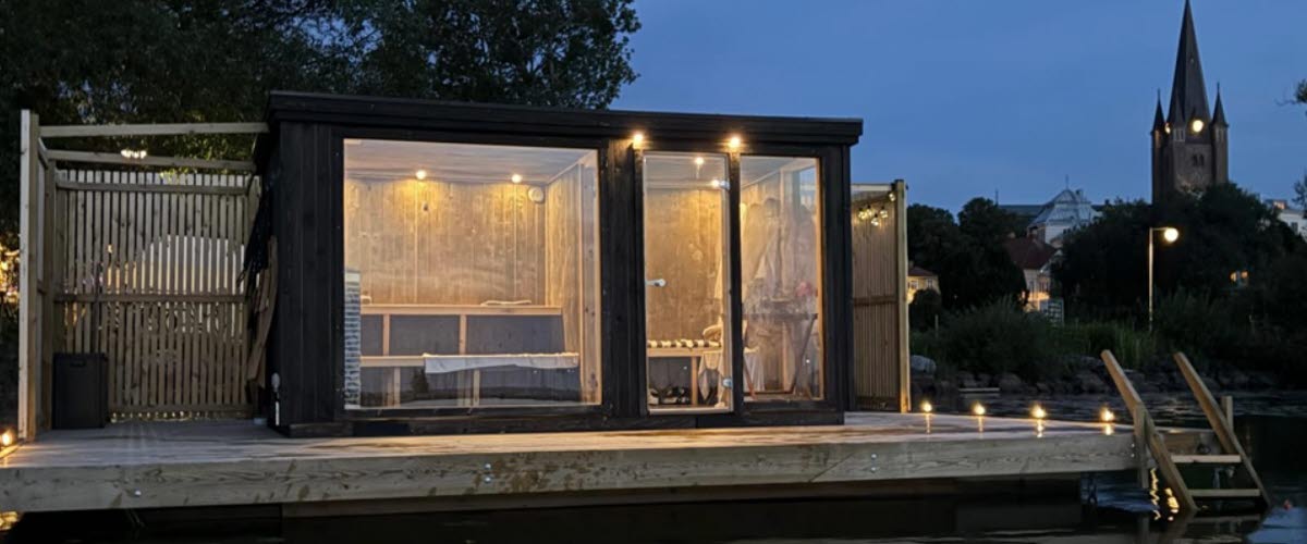 Bastuflotte i skymningen belyst innuti med en spegling i vattnet framför och siluetten av  domkyrkan i bakgrunden.