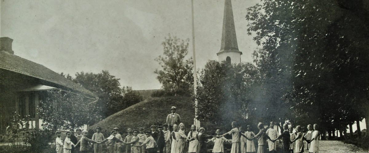 Äldre svartvitt foto som föreställer en skolklass där barnen står i ring och håller varandra i händerna. Bakom dem ser man Kung Ranes hög och Flistad kyrka. 