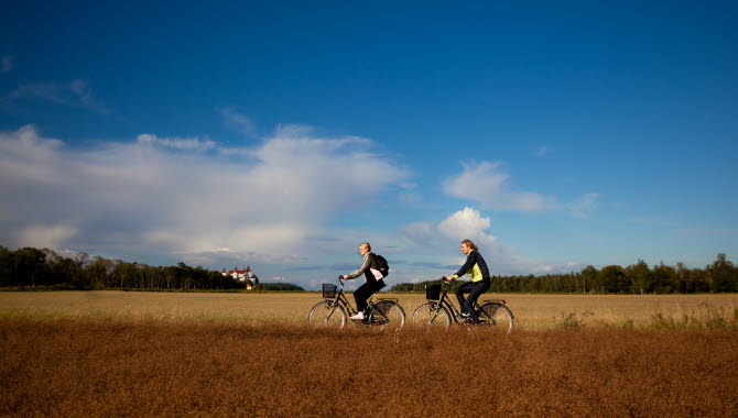 Two people rush forward on their respective bikes between open fields. Far away in the background, the white Läckö Castle can be seen.