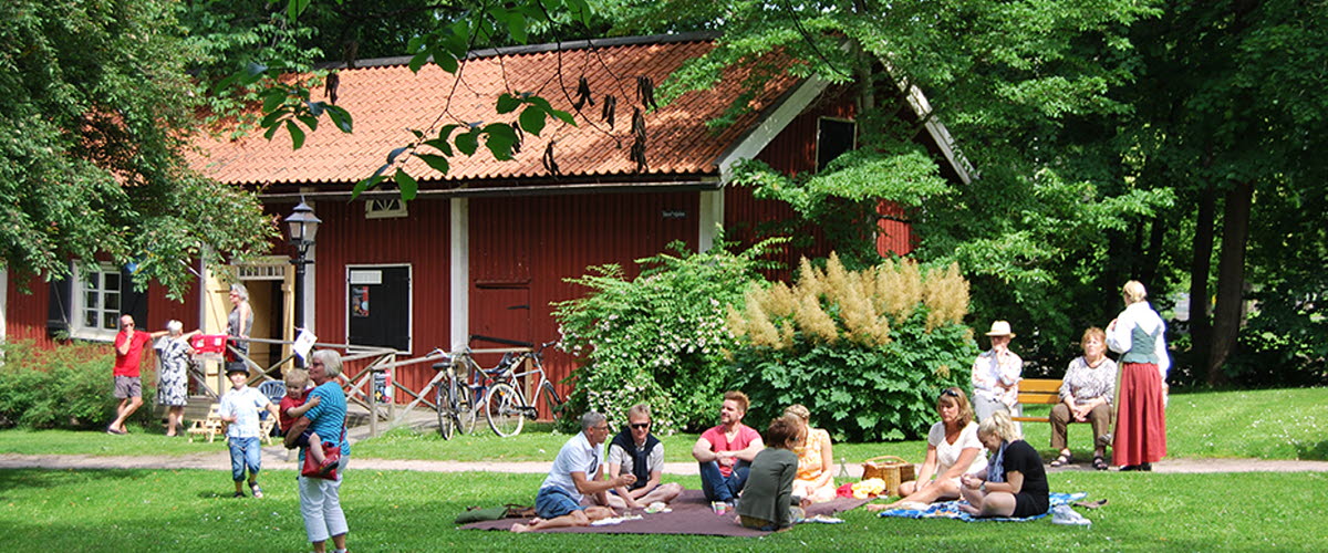 Personer som sitter i gröngräset och fikar och personer som går på Turbinhusön, som är en park med vackra gamla hus i Tidaholm.