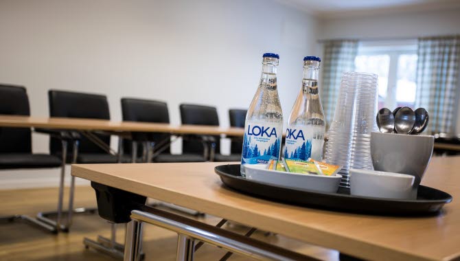 Bilden är tagen i en konferenslokal och i förgrunden syns en bricka med glas, skedar, frukt och mineralvatten. 