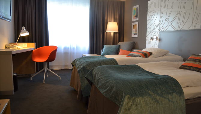 Två sängar med turkosa överkast står intill varandra inne på ett hotellrum. Framför sängar står även ett skrivbord med en orange stol.