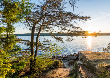 En bild på solnedgång över en stor sjö. I förgrunden syns träd och ien klippa.