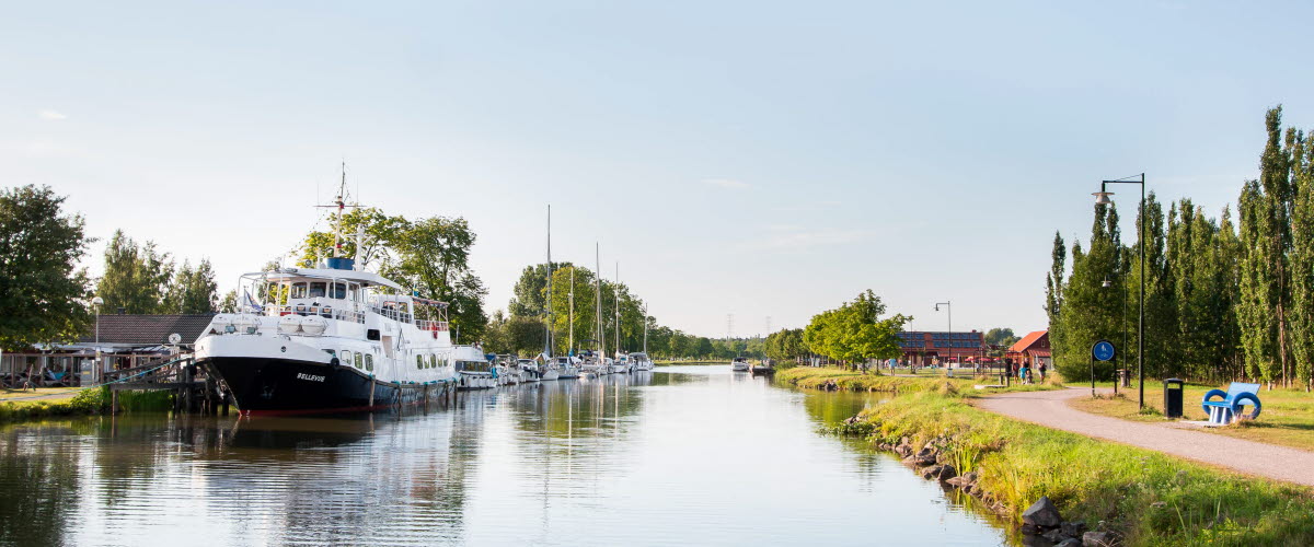 En stor vit passagerarbåt ligger vid Töreboda gästhamns brygg. Göta kanal syns och en promenadväg längs kanalen.
