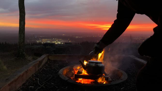 En kvinna fixar kaffet över öppen eld i soluppgången med utsikt över staden Skövde.