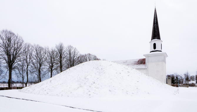 En stor gravhög täckt med snö. I bakgrunden skymtar taket och tornet på en kyrka.