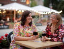 Two women in floral dresses sit at a table in an outdoor restaurant. In the background you can see more outdoor seating with string lights.