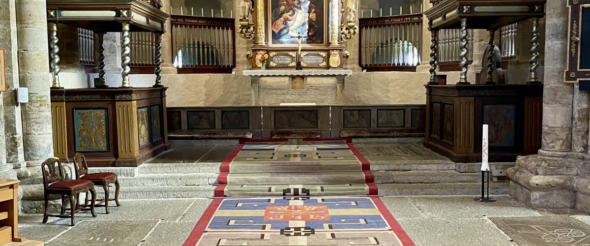 En vävd kormatta i ånga olika färger. Mattan ligger i en kyrka. På mattan står det Agda Österberg 1951