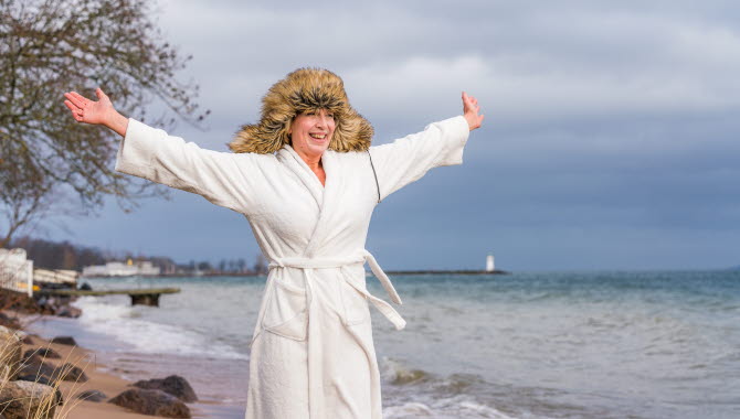 Kvinna står på strand i Hjo mitt i vintern, i morgonrock och pälsmössa.