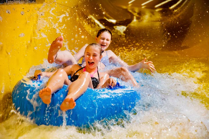 Två superglada tjejer åker på en blå badring i en gul vattenrutschkana så att vattnet stänker.