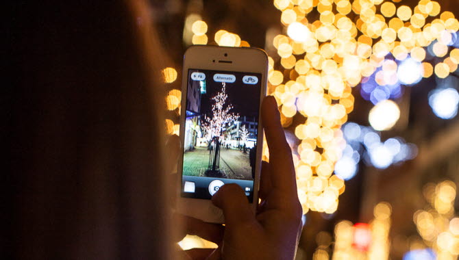 En person med långt hår håller upp en vit mobiltelefon och tar foto på ett träd med ljusslingor. Personen befinner sig utomhus och det är mörkt.