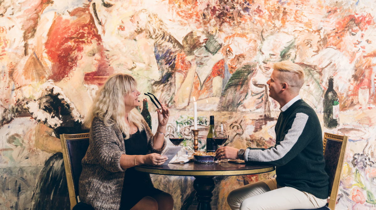 Två personer äter middag. Deras bord står framför ett konstverk som tar upp en hel vägg. Konstverket går i rödbrända toner.