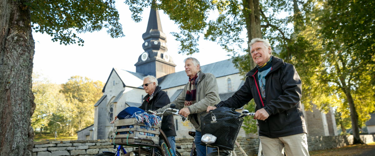 Tre män kommer gåendes och leder sina cyklar längs en stenmur. I bakgrunden ser man Varnhems klosterkyrka. 