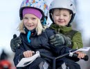 A girl with a colorful hat and helmet rides a snowracer with a boy sitting behind her wearing a green jacket and white helmet.