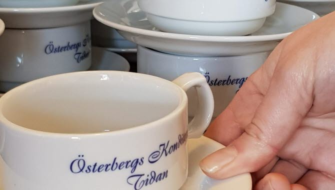 En hand lyfter en vit kopp med den snirkliga blå texten Österbergs Konditori Tidan