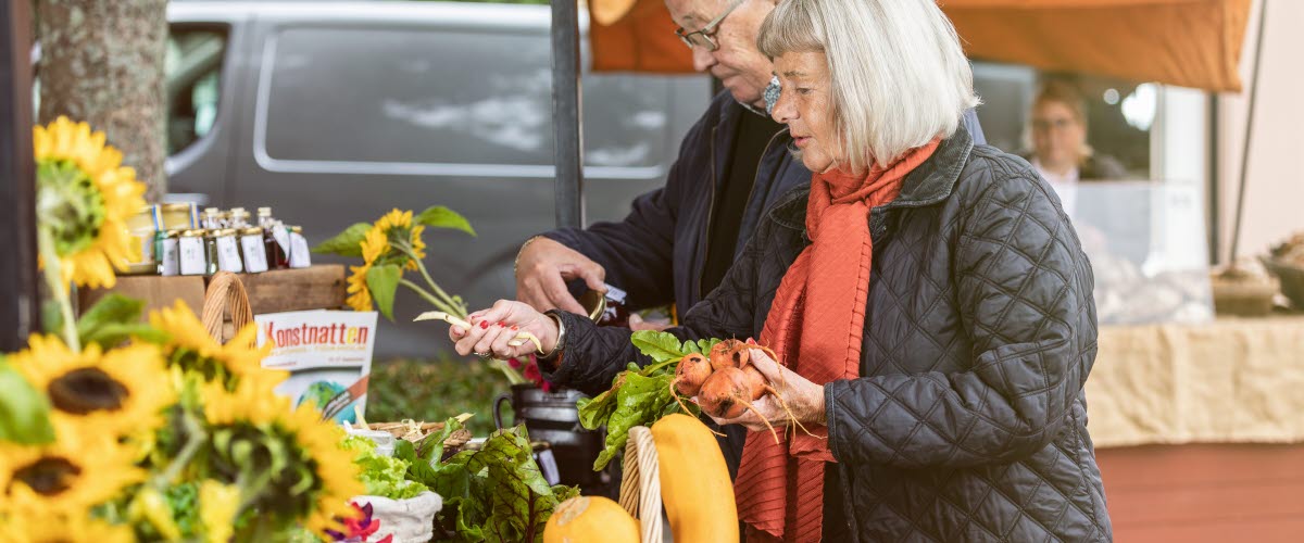 Man och kvinna som står och kikar på olika grönsaker på en marknad utomhus.