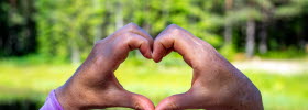 Two hands forming a heart. In the background, a lake and forest are visible.