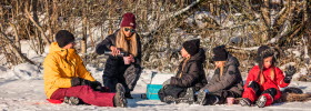 Man och kvinna och tre barn sitter ute i snön på filt och fikar.