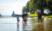 A family is wading in the water. The sun is shining and in the background you can se some trees an an old light house.