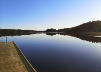 En bild på en blå sjö och en lika blå himmel. I bakgrunden skymtas en mörk skog. I vattnet ligger en brygga av trä.