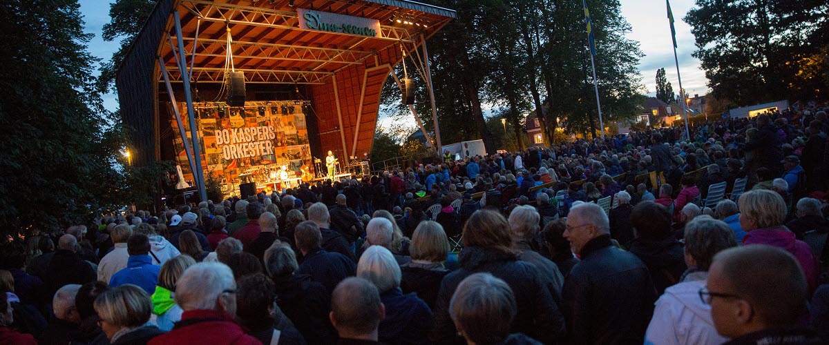 Konsert på utomhusscen i Stadsträdgården i Lidköping. Scenen lyser upp och framför står ett hav av människor.