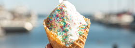 A hand is holding a soft serve ice cream cone with sprinkles on top. In the background, water is glimpsed.